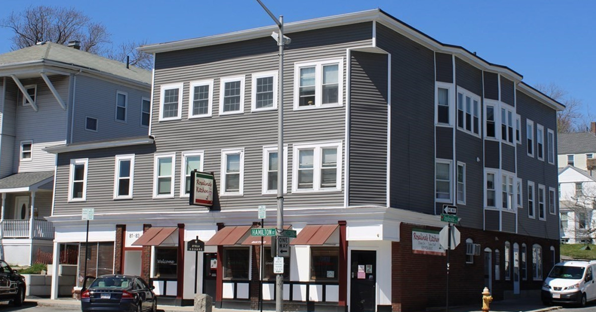 Large three story corner building with commercial and residential units, gray siding with white trim and red awnings out front